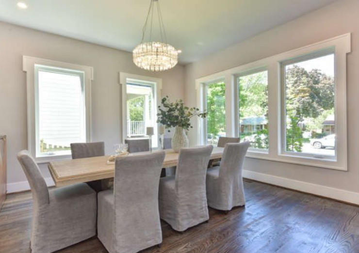 Bright dining room with a long wooden table and eight gray slipcovered chairs under a crystal chandelier, large windows bringing in natural light and greenery outside.