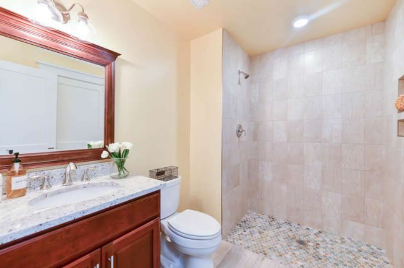 Bathroom with a dark wooden vanity, marble countertop, and framed mirror; modern fixtures, pump soap bottle, and a vase of flowers beside a toilet. A large tiled walk-in shower with a pebble mosaic floor is visible to the right.