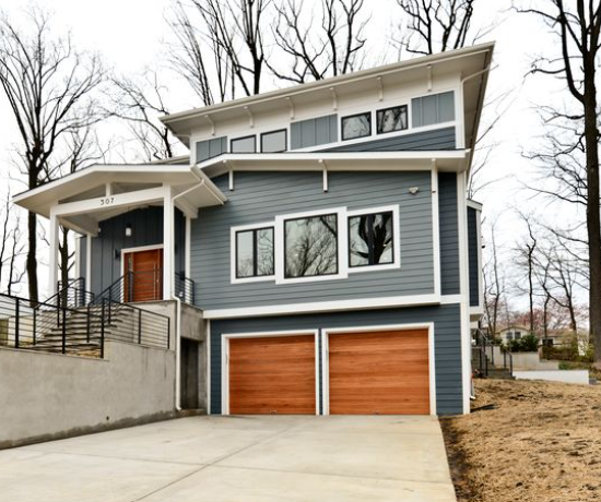 Two-story blue house with white trim and a double-car garage with wooden doors, a concrete driveway, and leafless trees behind.